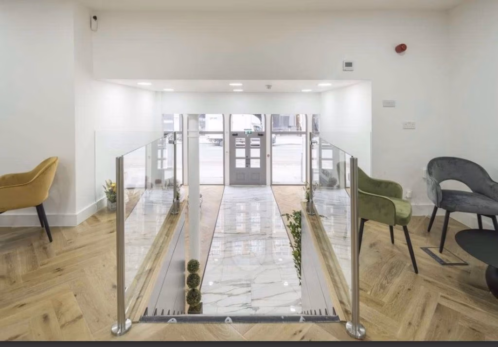 Bright lobby with a glass-railed stair/step down leading to glass double doors at the entrance; marble floor at lower level and wood herringbone flooring around; colorful lounge chairs along the walls.